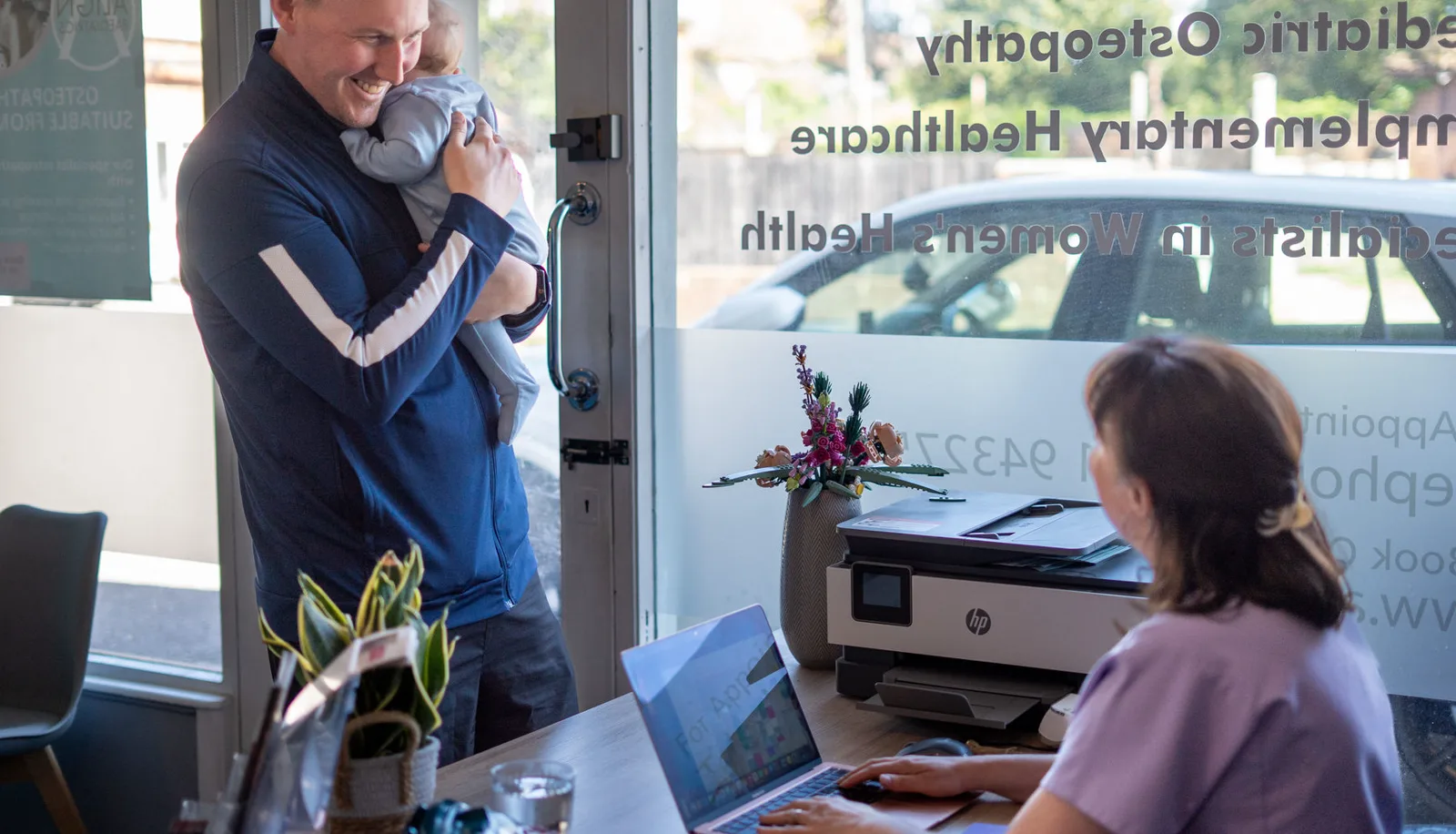 A father arriving at the Align Osteopathy reception with a baby and a toddler, the receptionist at her desk